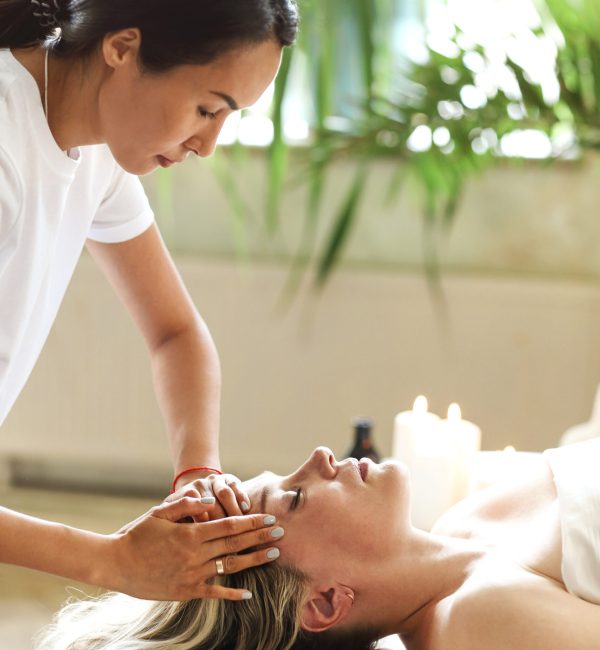 Side view of ethnic female therapist massaging face of middle age woman lying on massage table in spa salon